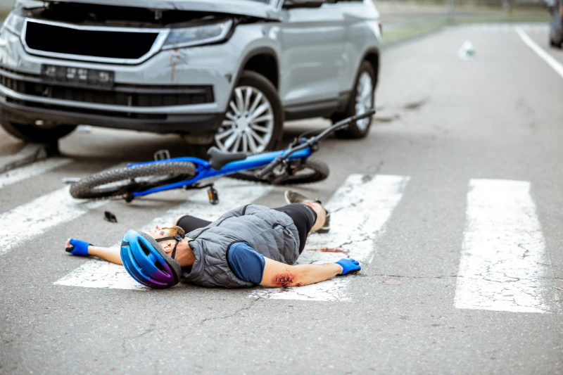 A person in a blue shirt lying on the ground next to a fallen bicycle after an accident, with another person kneeling by their side to help.